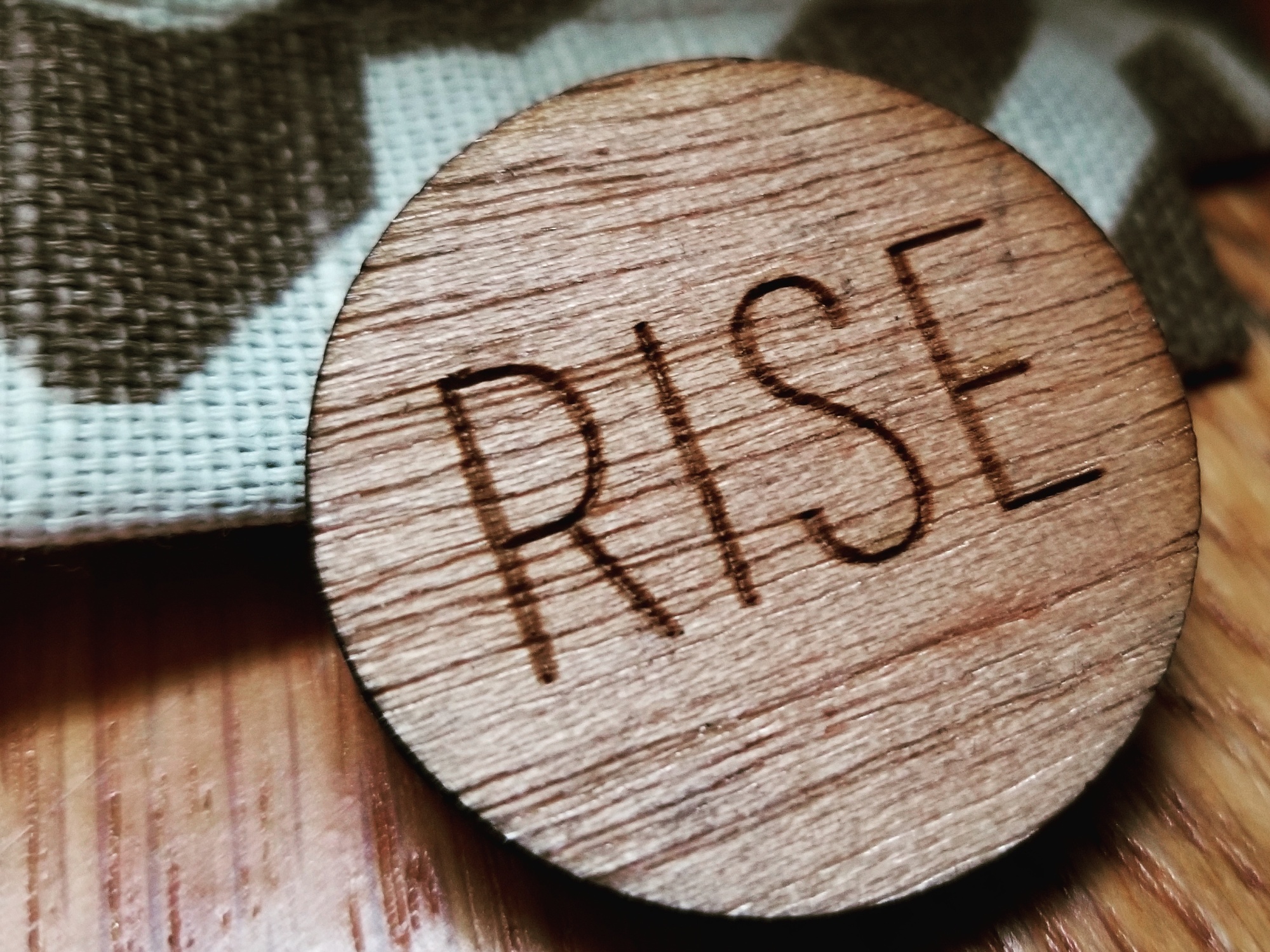 Photo of a brown wooden pin atop a brown and cream patterned textile. The pin says, "Rise."